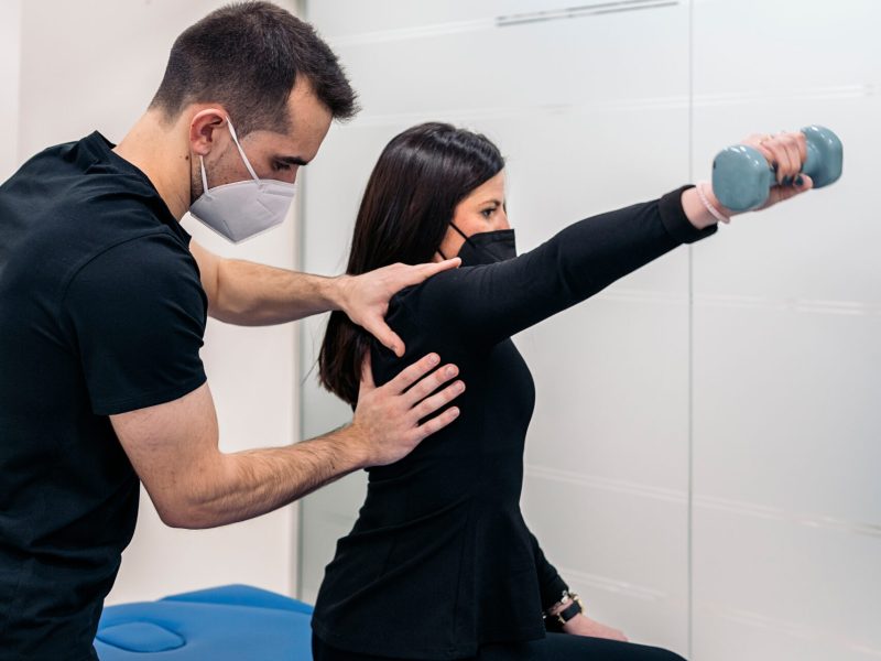 Male physiotherapist wearing face mask working with young woman in physiotherapy clinic.