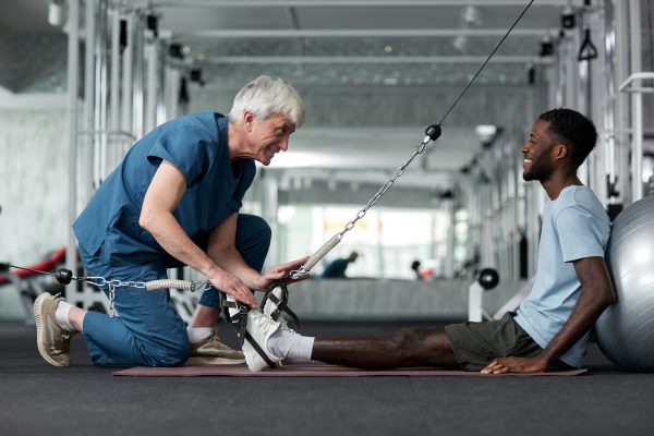 Portrait of smiling senior therapist helping young man with rehabilitation exercises in gym, copy space