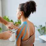 Close up rehabilitation therapist putting k tape on shoulder of young African American woman in physiotherapy clinic